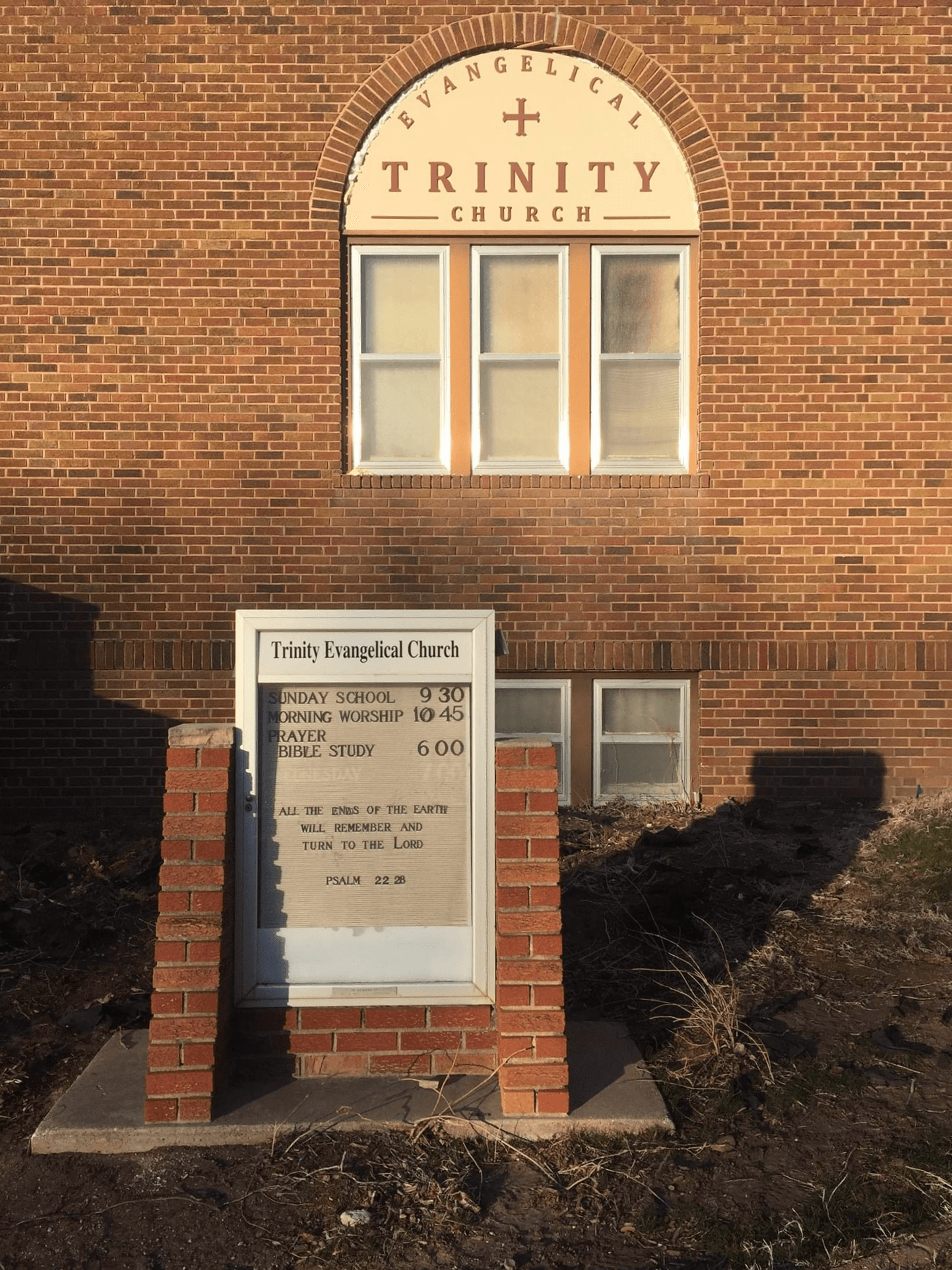 The church sign board mounted on the brick wall, listing Sunday School at 9:30 and Morning Worship at 10:45, beneath the arched 'Trinity Church' lettering.